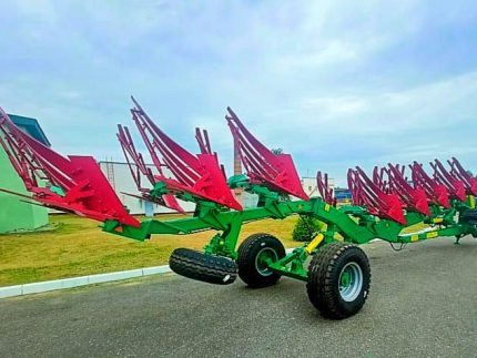 Сельскохозяйственный многокорпусный плуг с красными отвальными корпусами и зелёной рамой на колёсах на асфальтированной площадке